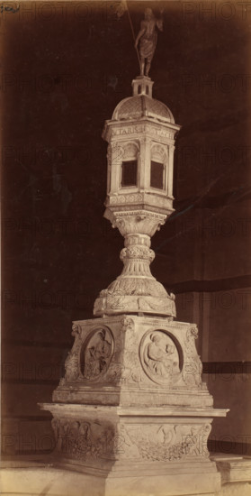 Tabernacle of St. Dominic's Basilica in Siena with Christ and the four evangelists, photograph, name: 'P. Lombardi Fotografo Siena', photographer: Paolo Lombardi, Siena, c. 1875 - c. 1900, Paolo Lombardi, photographer, Tuscany, paper, pasteboard, albumen print, height 266 mm x width 136 mm, Christ, tabernacle, altar, the four evangelists, other themes, A tall ornate stone monument stands against a dark background, featuring a square base with carved circular relief panels on each visible side depicting seated human figures. Above the base a fluted pedestal rises with elaborate floral and scrollwork carving, topped by a multi-sided lantern-like structure with arched window openings and decorative molding. The lantern is capped by a domed roof with textured patterning and a small tower supporting a statue of a standing human figure holding a long object. The surface shows aged stone tones and sculptural detail throughout.