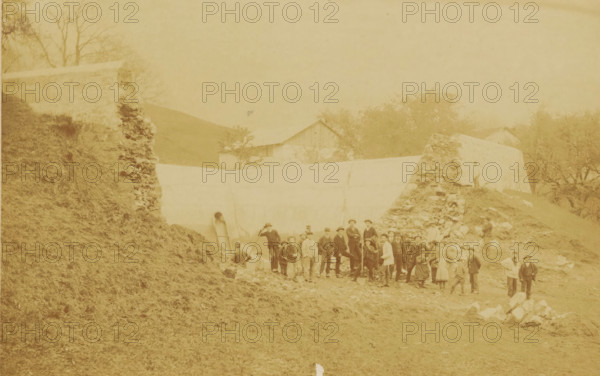 Workers at a construction site, photograph, photographer: Edouard Boulland, Vevey, c. 1880 - c. 1900, pasteboard, paper, albumen print, albumendruk, height 332 mm x width 402 mm, building-site, anonymous historical persons depicted in a group, in a group-portrait, A faded sepia photograph showing a group of people standing and posing on rough, rocky ground in front of a low wall and a pile of stones, with a sloped grassy embankment on the left and trees and simple gabled buildings in the background; the figures include men, women, and children wearing period clothing and hats, some holding tools or leaning on walking sticks, arranged in loose clusters across the central foreground, while a couple of individuals stand apart near the right edge and another person stands on the stone pile.