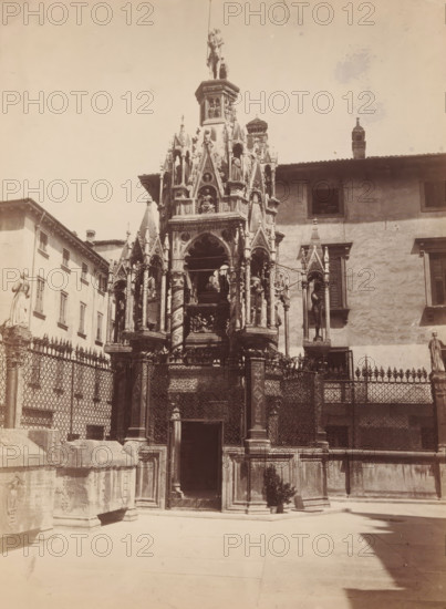 Tomb decorated with towers and sculptures, presumably in Italy, photograph, Europe, c. 1875 - c. 1900, photographer, paper, pasteboard, albumen print, height 299 mm x width 220 mm, monument, statue, grave-building, monumental tomb, A tall ornate stone monument with pointed arches and carved figures rises in front of a plastered building with shuttered windows, flanked by decorative iron fencing and stone posts; the monument features multiple statues set in niches, spiral columns, intricate relief work, and a small doorway at its base, while statues stand atop nearby pedestals and a paved open area stretches in the foreground.