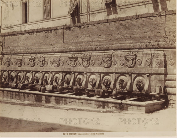 ANCONA Fontana delle Tredici Cannelle, Fountain of Calamo in Ancona, photograph, number: '4273.', Ancona, c. 1875 - c. 1900, photographer, Marche, paper, pasteboard, albumen print, albumendruk, height 189 mm x width 239 mm, ornamental fountain, A stone wall with decorative carved reliefs runs across the image, above a row of circular basins each set beneath a sculpted lion head spout; clay jars and wooden barrels sit in the basins and on the ledge below, while water trickles from the spouts into the basins; above the wall there is a building facade with shuttered windows and an open doorway, and the wall shows weathering and patches of vegetation; the foreground is a paved stone plaza with light and shadow across its surface.