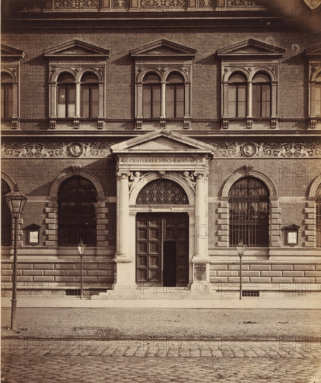 Façade of the Österreichisches Museum für angewandte Kunst in Vienna, Oesterr. Museum for Art and Industry, DETAIL', photograph, number: 'Nr. 64.', Vienna, publisher: Ludwig Niernberger, c. 1875 - c. 1900, Ludwig Niernberger, photographer, publisher, Lower Austria, paper, pasteboard, albumen print, height 233 mm x width 193 mm, column, pillar, architecture, façade (of house or building), A symmetrical stone and brick building façade with a central ornate entrance framed by columns and a triangular pediment, flanked by arched windows with decorative keystones and iron grilles, a frieze of sculpted relief running above the entrance and windows, pairs of tall rectangular windows with rounded tops and paired columns on the upper level, stone steps leading to a large paneled wooden door set within an arched opening with an ornate grille at the top, two cast iron street lamps mounted on poles in front of the building, a paved sidewalk and a cobbled street in the foreground, and decorative ar...