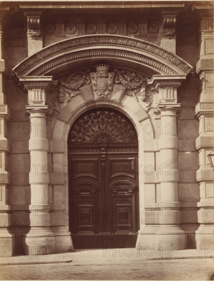 Palais Larisch, PORTAL', Gate of Palais Larisch-Mönnich in Vienna, photograph, number: 'Nr. 23.', Vienna, publisher: Ludwig Niernberger, c. 1875 - c. 1900, Ludwig Niernberger, photographer, publisher, Lower Austria, paper, pasteboard, albumen print, height 246 mm x width 190 mm, flowers, ornament, coat of arms (as symbol of the state, etc.), gate, entrance, A stone building facade with a large ornate arched entrance centered between two fluted columns that rest on rusticated bases, a carved semicircular fanlight with swirling ironwork sits above tall dark wooden double doors that feature raised panels, the arch is decorated with layered moldings and a carved crest flanked by floral garlands and ribbons, the entablature above shows rosette and bead motifs and supporting brackets, the surrounding masonry displays smooth ashlar blocks and textured rustication, and a cobblestone street runs along the base of the building.
