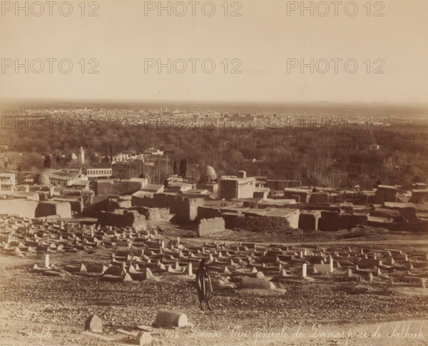 View of Damascus from Salhieh, Damas - Vue générale de Damas prize de Salhieh, photograph, number: '402.', name: 'JAG DE LEUR Représentant Huigenspark, 40b. THE HAGUE.', photographer: Maison Bonfils, Damascus, c. 1875 - c. 1900, Ottoman Empire, paper, pasteboard, albumen print, height 217 mm x width 270 mm, city-view, and landscape with man-made constructions, A wide sepia-toned landscape showing a foreground filled with numerous low rectangular and mound-shaped stone structures arranged in rows on uneven ground, with a solitary person standing among them wearing loose clothing and a head covering, a midground of clustered flat-roofed buildings and domed structures interspersed with trees, and a distant city skyline with many small buildings and taller spires on the horizon beneath a vast pale sky.