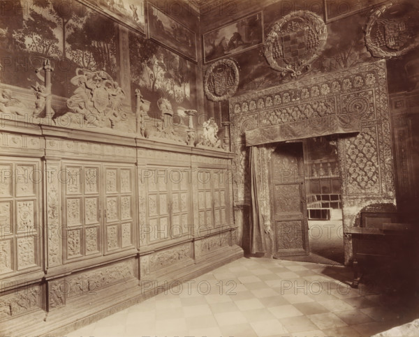 TOLÈDE. LA CATHÉDRALE, Entrance hall to the chapter house of the Cathedral of Toledo, photograph, number: '1396', inscription: 'Spain', Toledo Cathedral, c. 1875 - c. 1900, photographer, Toledo, Castilla-La Mancha, paper, pasteboard, albumen print, height 220 mm x width 277 mm, vestibule, hall, interior of church, painting, applied arts, art (relief, sculpture), A sepia-toned interior view showing an ornately carved wooden wall with rectangular panels and decorative reliefs, a tiled floor in a checkerboard pattern, and an open carved door partially covered by a patterned drape leading into an adjacent room; the upper walls display detailed sculptural friezes with figures, vases, and cherubic forms, painted murals of trees and foliage, framed landscape paintings near the ceiling, and large circular wreath-like medallions with heraldic designs, while the doorway surround and adjacent wall feature dense geometric and floral carved patterns and textiles.