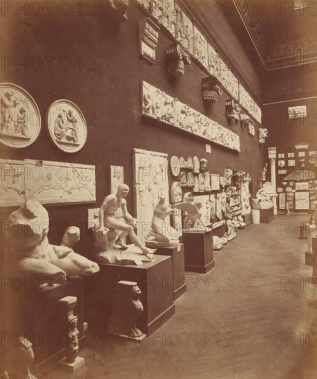 Arrangement of plaster casts (presumably) in the Musée des monuments français in Paris, photograph, Paris, c. 1875 - c. 1900, photographer, paper, pasteboard, albumen print, height 242 mm x width 203 mm, sculpture, permanent exhibition, museum, model (, production), A long gallery wall filled with sculptural reliefs and three-dimensional sculptures arranged on pedestals and shelves, featuring circular relief plaques, rectangular friezes, carved architectural fragments, and busts; several seated and reclining marble figures are displayed in the foreground on blocky stands, including a partially intact torso, a seated nude figure, and a crouching animal-like sculpture; the wall above shows a horizontal band of carved panels and ornate bracket-like elements, while smaller decorative plaques and sculpted heads occupy the mid and lower sections; the floor is wood with a herringbone pattern and the ceiling has decorative architectural detailing visible at the top right.