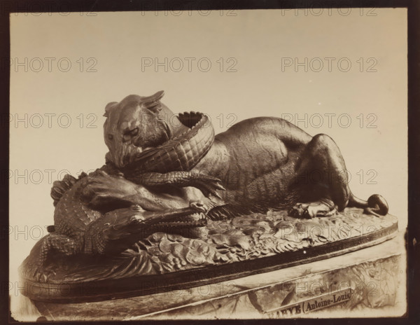 Image of a tiger devouring an alligator, by A. Barye, photograph, inscription: 'XI pl 9', c. 1875 - c. 1900, photographer, paper, pasteboard, albumen print, height 227 mm x width 286 mm, sculpture, beasts of prey, predatory animals: tiger, crocodiles: alligator, A sculpted scene showing a reclining big cat with textured fur and a lowered head, its neck encircled by a coiled reptile with scaly skin and visible claws, both animals resting on an ornate base with carved foliage and a rounded edge, photographed against a plain light background.