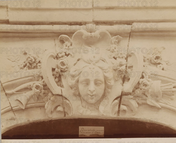 XVIIth century - Bas-relief provenant d'une maison à Caen., Bas-relief with a woman's face, from a house in Caen, photograph, name: 'A. GIRAUDON EDITOR 15. R. BONAPARTE. PARIS', inscription: 'pl 19', number: 'T 424', photographer: Adolphe Giraudon, Paris, c. 1875 - c. 1900, paper, pasteboard, albumen print, height 196 mm x width 241 mm, flowers, ornament, mask, mascaron, sculpture (relief, sculpture), A carved stone relief showing a stylized human face with closed eyes and a slight smile, framed by flowing hair and ornamental scrolls, surrounded by carved flowers and leaves, with curved architectural molding above and beneath and visible cracks and weathering on the surface.