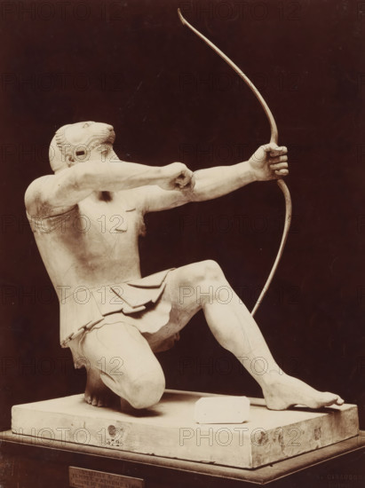 Art grec - Archer. Fronton du temple d'Ergine, Munich, Sculpture of an archer with a lion's hood, from the pediment of the temple at Aegina, photograph, number: 'T 572', name: 'A. GIRAUDON EDITOR 15. R. BONAPARTE. PARIS', inscription: 'pl 4 style k', photographer: Adolphe Giraudon, Paris, c. 1875 - c. 1900, paper, pasteboard, albumen print, height 253 mm x width 186 mm, sculpture, archer's weapons: bow and arrow, lion's head, ornament, shooting with bow and arrow, archery (, games of skill), A pale stone statue of a kneeling male figure wearing a short skirt and a headpiece shaped like an animal's head is shown from the side against a dark background, the figure holding a long curved bow in one hand while the other arm is drawn back as if pulling an invisible string, with visible musculature and detailed carving on the limbs and garments, the statue mounted on a rectangular base with a small rectangular block resting on the base near the figure's foot.