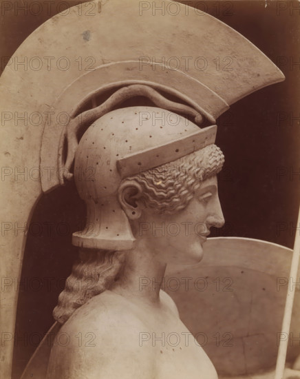 Head of the statue of Pallas Athena, from the pediment of the temple at Egina, Art grec - Tête d'Athene. Fronton du temple d'Egine. Munich, photograph, number: 'T 570 A', inscription: 'pl 4 style k', name: 'A. GIRAUDON EDITOR 15. R. BONAPARTE. PARIS', photographer: Adolphe Giraudon, Paris, c. 1875 - c. 1900, paper, pasteboard, albumen print, height 257 mm x width 199 mm, sculpture, helmet, head (human), (story of) Minerva (Pallas, Athena), A close-up profile of a pale stone sculpture showing a head wearing a large crested helmet with a curved plume, a textured band above the brow, and small perforations across the dome, the face turned slightly forward with a straight nose and closed lips, wavy hair peeking from beneath the helmet and gathered into a long braided lock down the back, a smooth shoulder and upper torso visible, and broad rounded armor or shield elements framing the head against a dark background.