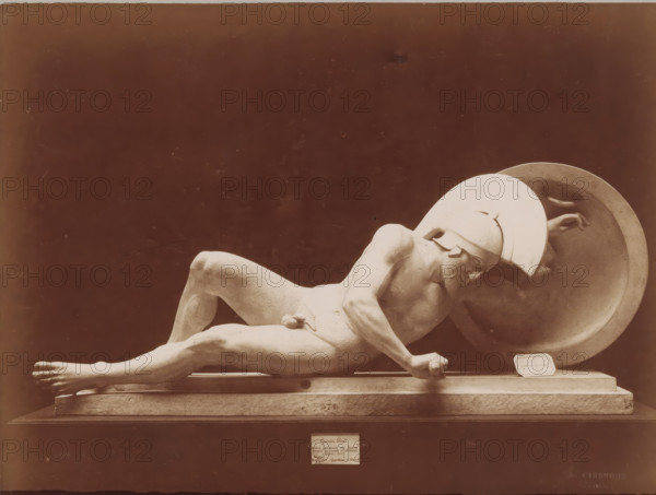 Sculpture of a wounded soldier, from the pediment of the temple at Aegina, Art grec - Guerrier blessé. Fronton du temple d'Egine. Munich, photograph, name: 'A. GIRAUDON EDITOR 15. R. BONAPARTE. PARIS', inscription: 'pl 4 style k', number: 'T 571', photographer: Adolphe Giraudon, publisher: Adolphe Giraudon, Paris, c. 1875 - c. 1900, Adolphe Giraudon, photographer, publisher, paper, pasteboard, albumen print, albumendruk, height 199 mm x width 255 mm, sculpture, helmet, lying figure, private soldier, protective weapons: shield, A reclining stone sculpture of a male figure wearing a helmet and holding a large round shield rests on a rectangular base against a dark background, the figure shown in profile with a bent arm gripping a short cylindrical object, a muscular torso and extended legs with visible toes, the helmeted head tilted forward and a hand visible near the shield's rim, the sculpture rendered in a pale, weathered stone with subtle surface texture and shadowing.
