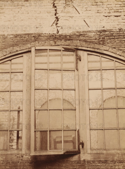 Detail of a window section at the Binnenhof during the restoration, photograph, annotation: 'Courtyard between the Knight's Hall and the Roll Room, photo taken there', annotation: 'Count's Halls, Restoration of the courtyard between the Knight's Hall and the Roll Room, April 1899, The Hague', Binnenhof, 1899, photographer, The Hague, Attributed to, paper, pasteboard, albumen print, height 224 mm x width 167 mm, closed window, A large arched multi-pane window set into a brick wall, with wooden frames and muntins dividing the glass into rectangular sections, some panes showing reflections and textured surfaces behind them, a vertical wooden post between the window sections, metal hinges and a latch attached to the frames, weathered brickwork above and below the window with visible mortar lines and a horizontal stone lintel, areas of chipped and worn paint and surface irregularities on both wood and brick.
