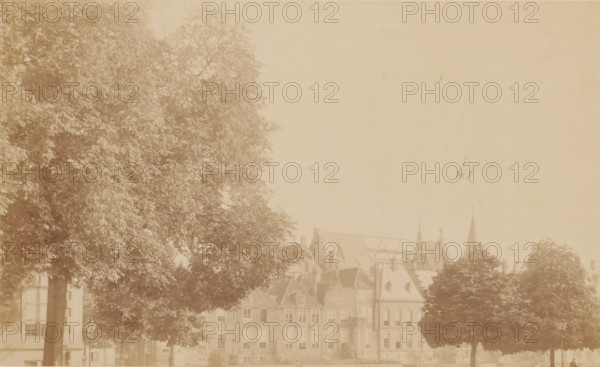 View of the Binnenhof with the Ridderzaal during restoration, photograph, annotation: 'The Hague, the Ridderzaal and the buildings of the Binnenhof seen from the Tournoiveld, July 1, 1902', annotation: 'Vijverberg, Viewed from Tournoiveld towards the buildings of the Ministry of the Interior etc. The roof (under restoration) of the Ridderzaal rises above it, 1902, The Hague', Tournooiveld, photographer, The Hague, Attributed to, paper, pasteboard, albumen print, height 139 mm x width 230 mm, national government, Binnenhof, A faded sepia-toned scene showing the upper portions of multi-story buildings with steep roofs and chimneys partially obscured by large leafy trees, with a tall spire and roof trusses visible above the rooftops, broad tree canopies dominating the left and right sides, a lamp post near the base of a tree, and an open pale sky occupying much of the upper area.