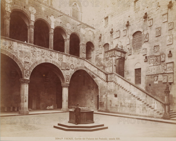 Courtyard of the Palazzo del Podestà in Florence, Italy, Cortile del Palazzo del Podestà, secolo - 1881, Giacomo Brogi, photographer, Florence, Tuscany, Attributed to, pasteboard, paper, albumen print, height 316 mm x width 422 mm, courtyard, palace, Palazzo del Podestà (Florence), A stone courtyard with a central octagonal well on a raised platform and a man leaning on its edge, a wide stair with stone steps ascending along a wall decorated with numerous carved plaques and reliefs, arched openings supported by columns with decorated capitals, a second level balustrade carved with heraldic and figurative reliefs, grated windows set into the masonry, sculptural elements and decorative panels mounted on the walls, dark recessed archways beneath the arcade, and scattered stone artifacts visible in the shaded areas.