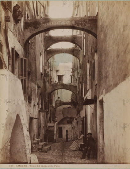 Street view with some people in San Remo, Italy, Vicolo del Giuoco della Palla, Sanremo, photograph, number: '8091.', collector's mark: 'VOSMAER', inscription: 'doubl', photographer: Carlo Brogi (possibly), San Remo, publisher: Edizioni Brogi, Florence, 1881 - 1900, Carlo Brogi, Edizioni Brogi, photographer, publisher, Tuscany, pasteboard, paper, albumen print, hand coloring, height 421 mm x width 316 mm, public road, peninsula, A narrow cobblestone alley flanked by tall, weathered buildings with shuttered windows and rough plastered walls, crossed overhead by a series of worn stone arches creating a tunnel-like perspective, with hanging laundry and small balconies visible along the upper levels, a street lamp suspended from one wall, stone steps and low ledges lining the sides, and several people present including two figures looking down from a high window and a seated woman in a long dress with another person nearby, plus a dog standing partway down the alley.