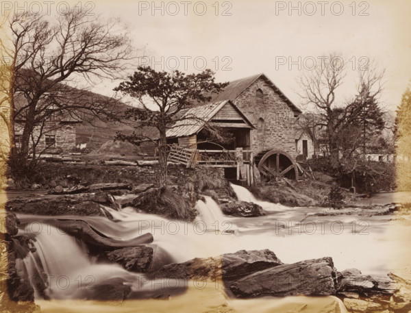Watermill at the Dochart in Killin, Mill on the Docharts, Killin, album page, photograph, This page is part of an album., number: '746, JV', c. 1875 - c. 1885, photographer, Stirling, paper, albumen print, height 298 mm x width 236 mm, watermill, A stone building with a pitched roof sits beside a flowing stream, a large wooden water wheel attached to its side and a wooden shelter extending from the structure; a tree with bare branches stands in front of the building and another tree with denser foliage is nearer the center, while fallen logs, wooden fences, and rough stone walls are scattered around the yard; cascading water flows over rocks in the foreground forming small waterfalls, and additional leafless trees and shrubs frame the scene on either side under a pale sky.