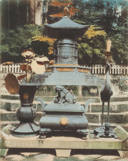 Tomb of Ieyasu, Nikko, photograph, annotation: '38. Tomb.', c. 1895 - c. 1915, photographer, Kanto, paper, pasteboard, salted paper print, Salt print, colored., height 264 mm x width 208 mm, height 329 mm x width 243 mm, A stone and metal shrine setup sits on tiered stone steps with a large ornate metal canopy structure centered toward the top, flanked by dense foliage and trees in the background; in front of the canopy a rectangular pedestal features decorative panels and a rounded top section, and on the lower platform a sculpted guardian lion rests atop a boxy incense burner with curved handles and a round ornament on its front; to the left a tall vase holds dried lotus pods and large round leaves, and to the right a tall, slender metal stand supports a slender candle above a turtle-shaped base; the scene shows weathered surfaces, patinaed metal, mossy stone, and a muted palette of greens, browns, and faded blue-gray tones.