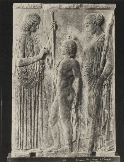 Demeter, Persephone and the child Triptolemos, Déméter, Perséphone & l'enfant Triptolème, photograph, number: '45.', Greece, c. 1895 - c. 1915, photographer, pasteboard, paper, collotype, photoprint, height 280 mm x width 218 mm, height 328 mm x width 239 mm, A carved stone relief showing three figures in profile standing close together against a rectangular panel, with two taller robed figures flanking a smaller nude figure in the center; the taller figures wear long draped garments with detailed vertical folds and hold staffs, one with wavy shoulder-length hair and the other with hair pulled back, while the central figure has short curly hair and a slender youthful body; the taller figures reach toward the central figure with one hand each, their garments showing layered folds and textured fabric, and the surface of the stone exhibits weathered texture and varying tones.