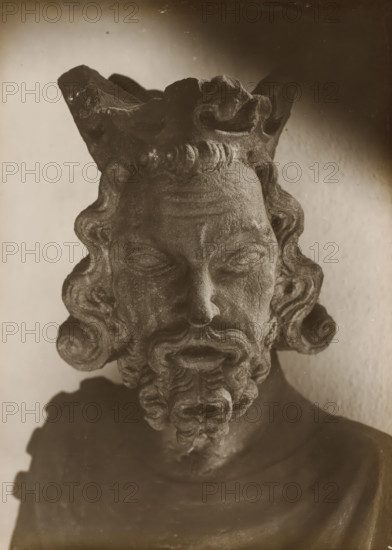 Sculpture in the cloister of Bamberg Cathedral, Cloister, Head, album page, photograph, This photo is part of an album., number: '148.', Bamberg, 1900 - c. 1920, photographer, Bavaria, paper, baryta paper, gelatin silver print, height 225 mm x width 164 mm, interior of church, Bamberg Cathedral, A close view of a weathered stone bust showing an aged face with closed eyes, a pronounced nose, curled hair framing the forehead and temples, a full curled beard with layered ringlets, and a textured crown-like headdress at the top, all rendered in monochrome tones against a softly lit plain background.