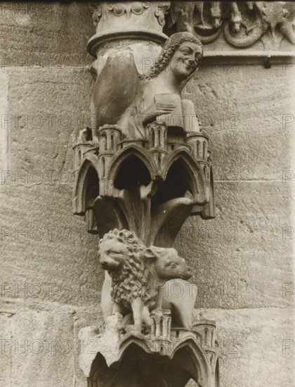 Adamspforte, Engel, Löwe, Ochse vN, Detail of the Fürstenportal in Bamberg Cathedral, album page, photograph, This photo is part of an album., inscription: 'Kunsthist. Seminar, Marburg (Lahn), Photogr. Abteilung, Vervielfältigung, untersagt.', number: '62', Bamberg, 1900 - c. 1920, photographer, Bavaria, paper, baryta paper, gelatin silver print, developing gelatin silver print, height 238 mm x width 181 mm, church (exterior), Bamberg Cathedral, A carved stone figure perched on an ornate bracket against a masonry wall, showing a smiling human face with curled hair and a hand holding a rectangular object, above a detailed canopy of pointed arches and small columns, with a sculpted lion head and an animal head partially visible below, all rendered in weathered stone tones and set against the textured stone blocks of the building.