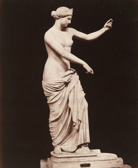 Venus of Capua, Museo di Napoli Venere, album page, photograph, This photo is part of an album., number: '7507', workshop stamp: 'Giorgio Sommer, Largo Vittoria, Napoli', photographer: Giorgio Sommer, Naples, 1874 - in or before 1879, paper, albumen print, height 250 mm x width 203 mm, sculpture, (story of) Venus (Aphrodite), A pale marble statue of a draped female figure is shown against a dark background, the figure standing on a stepped pedestal with one foot forward and a cloth wrapped around the lower body displaying deep folds and flowing drapery; the torso and arms are bare, one arm extended upward with the hand slightly cupped and the other arm angled downward with the fingers relaxed, the head turned in profile with hair pulled back into a low bun and a band around the head, and the surface of the sculpture shows subtle shadows and smooth contours.