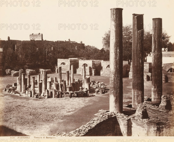 View of the ruin of the macellum of Pozzuoli, Pozzouli, Tempio di Serapide, album page, photograph, This photo is part of an album., number: '6197', workshop stamp: 'Giorgio Sommer, Largo Vittoria, Napoli', photographer: Giorgio Sommer, Pozzuoli, 1874 - in or before 1879, paper, albumen print, height 203 mm x width 252 mm, market-hall, ruin of a building, architecture, column, pillar, A wide view of stone ruins with many upright columns of varying heights and widths standing on a flat ground area, some columns grouped closely and others standing apart, several broken column fragments and scattered blocks lying near the bases, three very tall weathered columns dominating the right side with visible surface texture and irregular tops, low stone walls and foundation remnants framing parts of the site, a line of rectangular stone structures with arched openings set behind the columns, a dense row of trees and vegetation rising behind those structures, and a distant larger stone building wi...
