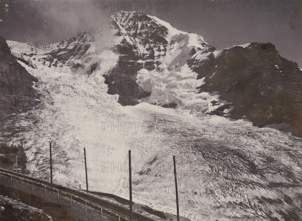Jungfraubahn und Mönch, View of the Mönch with the Jungfraubahn railway, photograph, This photo is part of a cover., inscription: 'Gebr. Wehrli. Kilchberg-Zurich. 3890. Jungfraubahn und Mönch.', Jungfraujoch, publisher: Gebrüder Wehrli, Kilchberg, 1912 - 1920, photographer, Alpen, Bernese Alps, paper, pasteboard, baryta paper, daylight collodion silver print, height 164 mm x width 218 mm, railway, train, mountain-top, peak, Alps, Mönch, A wide snowy mountain scene fills the image, showing a steep, rugged peak with layered rock faces and snow fields, a large glacier flowing down the central slope with crevasses and textured ice, darker rocky ridges flanking the snowy areas, and a railway track with tall vertical poles running along the lower foreground beside a narrow ledge.