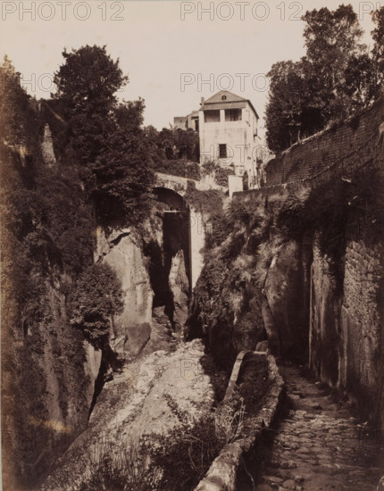 View of a gorge in Sorrento, Italy, Ravino di Sorrento, photograph, number: '137.', inscription: '(+) Sommer, MB, nakomen Diepr.', photographer: attributed to Giorgio Sommer, Sorrento, 1857 - 1914, Attributed to, pasteboard, paper, albumen print, height 327 mm x width 241 mm, landscape with bridge, viaduct or aqueduct, ravine, chasm, abyss, canyon, A sepia-toned scene showing a narrow rocky gorge with steep cliff faces covered in patches of vegetation, a cobbled footpath with a low stone railing running along the right edge, and stone steps descending beside a high retaining wall; at the top center a pale multi-story building with a tiled roof and a small balcony overlooks the gorge, flanked by trees and dense foliage, while a stone arch bridge spans a gap in the rock beneath the building.