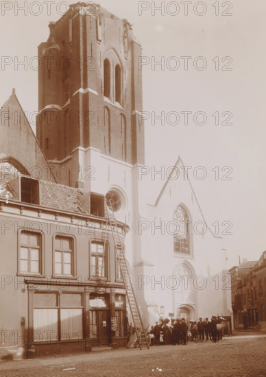 View of the Reformed Church in Vlissingen after the fire in 1911, photograph, annotation: 'JB van Bree, 713d, 2554', annotation: 'NH church in Vlissingen after the fire of September 6, 1911', Flushing, 1911, photographer, Attributed to, paper, pasteboard, daylight collodion silver print, height 211 mm x width 168 mm, church (exterior), street (city(-scape) with figures, staffage), A tall brick church tower with arched openings rises beside a lighter stone facade with large pointed arched windows; in front of the building a row of narrow townhouses with tiled roofs and dormer windows lines the street, one with a tall ladder leaning against it; a group of people stand near the church entrance, some wearing hats and a bicycle nearby; cobbled street in the foreground and additional buildings receding along the right side.