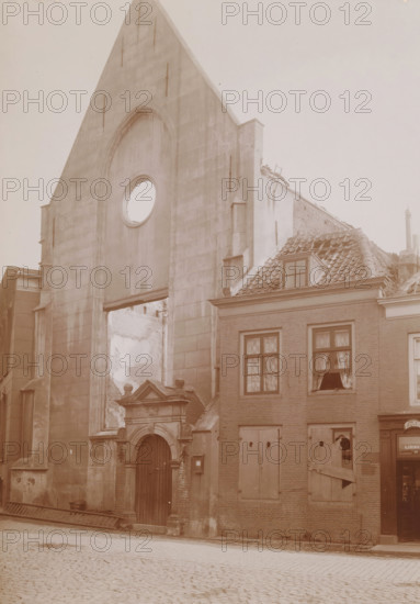 View of the Reformed Church in Vlissingen after the fire in 1911, photograph, annotation: 'NH church in Vlissingen after the fire of September 6. 1911', annotation: 'P. Louw, Ca. 1710', Flushing, 1911, photographer, Attributed to, paper, pasteboard, daylight collodion silver print, height 214 mm x width 152 mm, church (exterior), A tall stone façade with a pointed gable and a large circular opening sits next to a brick building with a tiled roof and a dormer window, the stone façade showing an arched recess and a decorated doorway with columns and a wooden door, while the adjacent brick building has shuttered and broken windows with curtains visible behind some panes and a boarded lower window, roof tiles appearing damaged and partially missing, rubble and exposed interior walls visible through gaps, and a cobblestone street in the foreground.