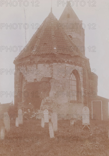 View of the Reformed Church of Wierum, photograph, annotation: 'Wierum (Fr) Ned. Rev. church, Sept: 1910, Westdongeradeel', Wierum, photographer: AJM Mulder (possibly), 1910, Attributed to, paper, pasteboard, daylight collodion silver print, height 220 mm x width 160 mm, churchyard, cemetery, church (exterior), A weathered brick building with a conical tiled roof and a taller square tower rising behind it, showing areas of flaking plaster and exposed brickwork; an arched window with multiple panes is set into the curved wall, and a plain wooden door is visible at ground level; in front of the building a grassy area contains numerous upright stone markers of varying shapes and sizes, some set in rows, with one taller marker standing near the door; the scene is muted and pale in tone, with a vertical streak of discoloration on the left side of the image.