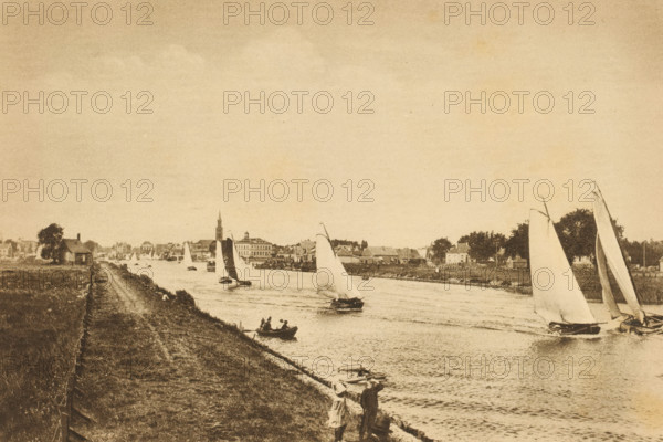 Sailing in North Holland, Sailing race on a waterway in North Holland, album page, photomechanical print, This print is part of an album., photographer: E. van Elfrinkhoff, North Holland, cliché maker: Nederlandsche Rotogravure Maatschappij, Leiden, in or before 1915, paper, rotogravure, height 161 mm x width 240 mm, sailing-ship, sailing-boat, river, A riverside scene in a muted sepia tone showing a stretch of grassy bank and a dirt path alongside a waterway with multiple sailboats of varying sizes on the water. Several boats have tall sails, some angled and some nearly vertical, and a small rowboat with figures is near the shore. Buildings and trees line the opposite bank, including a tower with a pointed top rising above nearby structures. Two people stand on the near bank looking toward the river, and scattered small boats are moored along the edge of the water.