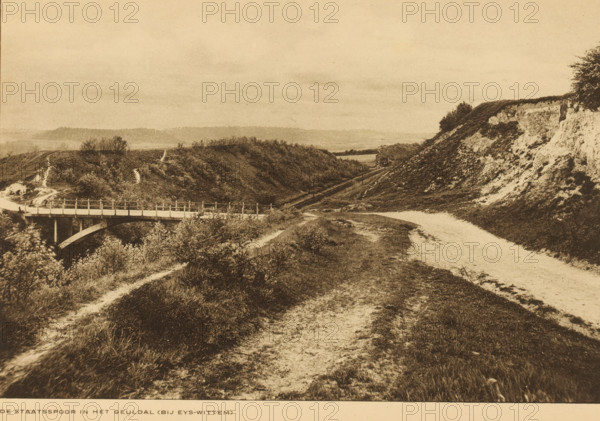 View of the Staatsspoorweg in the Geuldal near Eys, Limburg, The Staatsspoor in the Geuldal (near Eys-Wittem), album page, photomechanical print, This print is part of an album., cliché maker: Nederlandsche Rotogravure Maatschappij, Leiden, in or before 1915, photographer, paper, rotogravure, height 160 mm x width 239 mm, railway, train, low hill country, A sepia-toned landscape showing a dirt path curving alongside grassy slopes and a steep eroded hillside, with a bridge spanning a valley on the left and a small structure near the center-right at the base of the hills, scattered shrubs and trees on the ridges, and distant low hills under a wide sky.