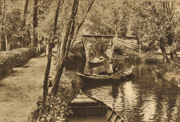 View of Giethoorn, Overijssel, Giethoorn, album page, photomechanical print, This print is part of an album., cliché maker: Nederlandsche Rotogravure Maatschappij, Leiden, in or before 1915, photographer, paper, rotogravure, height 161 mm x width 240 mm, prospect of village, silhouette of village, canals, waters (in city), gondola, A sepia-toned scene of a narrow river flanked by trees and vegetation with a wooden footpath running alongside one bank, several small rowboats on the water including one in the foreground tied to the bank, two men standing in separate boats holding long poles, a rustic wooden bridge arching over the river in the midground, dense foliage and tall trees on both sides, and reflections of the boats and trees visible in the water.