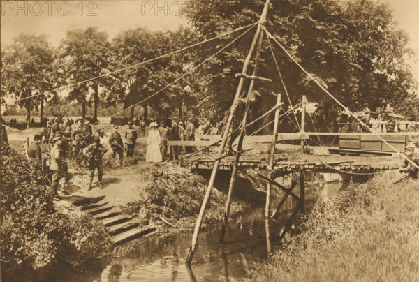 HM attends an exercise in troops crossing the water, Queen Wilhelmina attends a military training, in which soldiers must cross a river, album page, photomechanical print, This print is part of an album., photographer: NV Vereenigde Fotobureaux, Netherlands, cliché maker: Nederlandsche Rotogravure Maatschappij, Leiden, in or before 1915, paper, rotogravure, height 161 mm x width 240 mm, Person, Wilhelmina of the Netherlands, military training, queen, crossing a river, A sepia-toned outdoor scene showing a wooden footbridge over a narrow stream, supported by rough poles and ropes tied to a tall central mast, with a man climbing on the timber framework. Alongside the bridge and stream are grassy banks and a stepped dirt path leading down to the water. Groups of people stand near the bridge and under a line of large leafy trees in the background; some figures wear hats and one person in the foreground wears a light-colored dress. Ropes run from the mast to multiple anchor points across th...