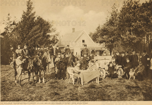 The division commander gives his orders through his staff, Commander during a meeting with his staff, album page, photomechanical print, This print is part of an album., Netherlands, cliché maker: Nederlandsche Rotogravure Maatschappij, Leiden, in or before 1915, photographer, paper, rotogravure, height 161 mm x width 240 mm, military training, Command, A sepia-toned outdoor scene showing a large group of uniformed men gathered in front of a wooden house with a steep roof and a small balcony, surrounded by tall trees; several men are mounted on horses, others stand or sit near a vintage open automobile, and a few men are seated at a table covered with a cloth while another man writes; some men wear peaked caps and others wear helmets, and a dog is sitting beside a seated man.