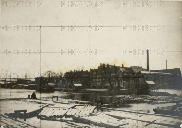 Westerdok under the snow in Amsterdam (former title), De Zandhoek in Amsterdam (former title), The Westerdok viewed towards the Zandhoek, photograph, The Westerdok and the Zandhoek under the snow in Amsterdam. Grid lines have been drawn on the photo in pencil., inscription: '84 x 158 1, 2', photographer: George Hendrik Breitner, Netherlands, 1902 - 1903, paper, pasteboard, gelatin silver print, developed gelatin silver print, height 361 mm x width 501 mm, height 380 mm x width 515 mm, harbour, Amsterdam, Westerdok, Zandhoek, A wide view of a waterfront scene with a wooden dock in the foreground showing planks and stacked logs partially covered in snow, a small bench or bollard near the water's edge, and a lone seated figure facing the water; across the water a dark silhouette of industrial and residential buildings lines the shore with rooftops and a tall chimney rising above them, a smaller slender smokestack further along, and leafless tree shapes interspersed among the structures; t...