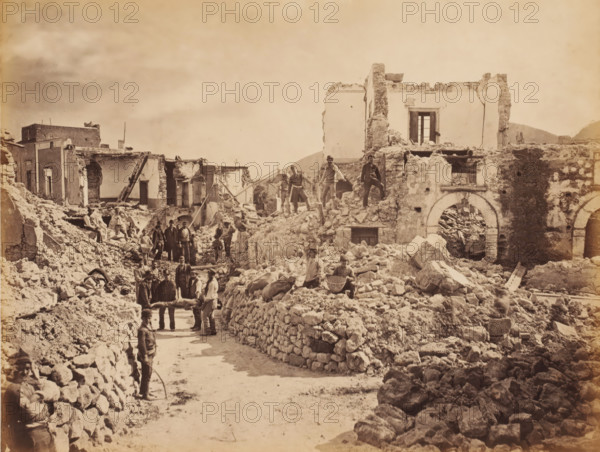 Destruction by an earthquake in southern Italy, photograph, annotation: 'f 30, -', inscription: 'Middle East 1890's', Italy (possibly), 1905 - 1908, photographer, Italy, pasteboard, paper, albumen print, albumendruk, height 188 mm x width 248 mm, height 238 mm x width 277 mm, ruin of a dwelling, house, castle, (carrying a person on a) litter, stretcher, consequences of an earthquake, Sicily, Calabria, A sepia-toned scene of ruined buildings and rubble filling a street, with crumbling stone walls, exposed interiors, and broken arches; several men stand or sit among the debris, some on higher piles of stone and others on the ground, a few appear to be carrying a stretcher-like object, and one man stands in the foreground holding a curved tool; scattered stones and broken masonry form makeshift low walls along the road, and remnants of doorways and window openings are visible in the damaged structures.