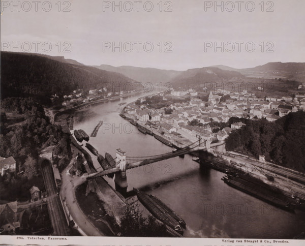 Panorama of the city of Decín (German: Tetschen-Bodenbach) on the Elbe, Czech Republic, photograph, Panorama of the city of Decín (German: Tetschen-Bodenbach) on the Elbe in the Czech Republic. Taken from a very high vantage point, placing the flow of the river and the location of the city in the center., inscription: 'Verlag von Stengel & Co., Dresden.', inscription: '3235. Tetschen, Panorama.', publisher: Stengel & Markert, Germany, c. 1890 - c. 1925, paper, photoaquatint, heliogravure, height 229 mm, width 284 mm, Severocesky Kraj, Elbe, Tetschen, A wide aerial view of a river winding through a town with a suspension bridge crossing the water, stone bridge towers at each end, and several long flat boats moored along the riverbanks. On one side of the river a dense forested hillside rises, dotted with small houses near the shore and a tunnel entrance visible at the base of the hill. On the opposite bank a compact cluster of buildings with pitched roofs and a church with a tower fill ...