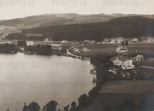 Titisee, Black Forest, Baden-Württemberg, Germany, photograph, The Black Forest lies on the Rhine and on the French border. On the northern shore of the lake, which covers 1.3 km², lies the spa town of the same name, nowadays part of the city of Titisee-Neustadt. Here we see Titisee from the South., number: '2313', inscription: 'Verlag der Neuen Photogr.Gesellschaft A.-G., Berlin-Stieglitz 1904', inscription: 'SCHWARZWALD. Titisee von Süden.', publisher: Photographische Gesellschaft Berlin, in or before 1904, paper, gelatin silver print, developing gelatin silver print, height 190 mm, width 242 mm, Titisee-Neustadt, A wide view of a lakeshore with calm water in the foreground and a shoreline dotted with trees, small docks, and boathouses, a curved road running along the shore past several standalone houses and larger multi-story buildings with pitched roofs, clusters of trees and manicured lawns around the buildings, open fields and farmhouses extending inland, and sloping forested hil...