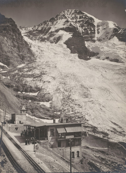 Jungfraubahn, Eigergletscher station, Mount Eiger in the background, Bernese Alps, Switzerland, photograph, Eigergletscher station with a 'Restaurant' sign, a few people on the platform, Switzerland. The railway was completed and opened in 1912., inscription: 'Gebr. Wehrli, Kilchberg - Zürich.', stamp: 'Algemeene, Boek-, Papier- en kunsthandel, GA KOTTMANN, 61 Valeriusstraat 61', inscription: '13878 Jungfraubahn Station Eigergletscher.', photographer: Gebrüder Wehrli, in or after 1912-08-01 - c. 1925, pasteboard, paper, gelatin silver print, developing gelatin silver print, height 216 mm, width 163 mm, A steep, snow-covered mountain slope fills the upper portion of the image, with jagged rock faces and layered ice formations visible on the peaks and ridges, contrasting light snow and darker exposed rock. Midway down the slope, the glacier shows crevassed textures and striations as it descends toward a rocky moraine. Near the base of the mountain, a small cluster of buildings sits on a ...