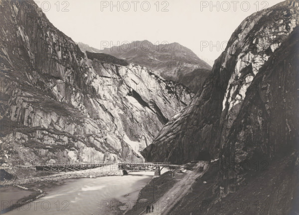 Tunnel 'Das Urnerloch' Schöllenen? and bridge over the Reuss, Gotthard Pass, Switzerland, photograph, To the right of the deep valley between steep rocks you can see the entrance to the 64-meter-long tunnel 'Urnerloch', built between 20. 09. 1707 and 15.08. 1708. A low bridge can be seen over the river Reuss., inscription: '2073. Das URNERLOCH.', inscription: 'Gebr. Wehrli, Kilchberg, Zurich.', photographer: Gebrüder Wehrli, c. 1900 - c. 1925, pasteboard, paper, gelatin silver print, developing gelatin silver print, height 161 mm, width 216 mm, A wide monochrome landscape showing steep rocky cliffs and craggy mountains with striated rock faces and lighter patches of exposed stone, a river flowing through the valley with a wooden bridge crossing it, a dirt road running alongside the river, and three small figures walking on the road near the foreground.