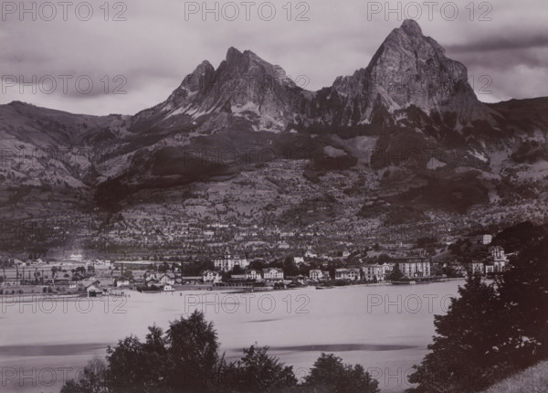 The place Brunnen and the two 'Myths' (mountain range), Switzerland, photograph, Brunnen is situated on Lake Lucerne in Central Switzerland. In the background, two striking mountain peaks - 'the two Myths' - can be seen., inscription: '1532. Brunnen & die Mythen', photographer: attributed to Gebrüder Wehrli, c. 1900 - c. 1920, Attributed to, paper, gelatin silver print, height 218 mm, width 165 mm, Brunnen, A wide grayscale landscape shows a large mountain range rising behind a tiered hillside dotted with trees and patches of lighter rock, with steep rocky peaks and ridges under a cloudy sky; below the slopes a built-up area with rows of multi-story buildings and smaller houses lines the shore of a calm body of water, where a small pier and a few boathouses project into the water; foreground trees frame the bottom edge of the image.