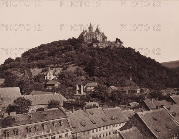 Wernigerode mit Schlossberg', Saxony-Anhalt, Germany, photograph, View of a castle located on a wooded mountain, with the roofs of the houses of Wernigerode in the foreground., inscription: 'E 11. Wernigerode m. Schlossberg.', c. 1885 - c. 1910, photographer, pasteboard, paper, albumen print, height 212 mm x width 274 mm, height 356 mm x width 455 mm, Wernigerode, A hill densely covered in trees rises above a town, topped by a large stone castle complex with multiple steep roofs, towers, turrets, and crenellated walls, while below the hill a cluster of houses and buildings with pitched roofs, dormer windows, and chimneys sit among trees and small clearings.
