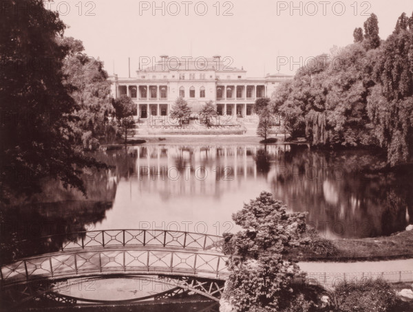 House in a park by a large pond, Frankfurt am Main ?, Germany, photograph, Germany., c. 1885 - c. 1895, photographer, pasteboard, paper, albumen print, height 202 mm x width 268 mm, height 508 mm x width 330 mm, A wide, symmetrical building with two stories of columned balconies and a central entrance sits at the far edge of a calm reflective body of water, its facade mirrored in the lake; flanking the building are dense clusters of tall trees and shrubs, with large trees framing the scene on both sides. In the foreground a decorative metal footbridge with geometric railing arches over a small inlet, and near the bridge a leafy bush and other low plants grow beside a gently sloping bank. The overall image has a sepia tone and soft, slightly faded contrast, with reflections of the building and surrounding trees visible on the water surface.