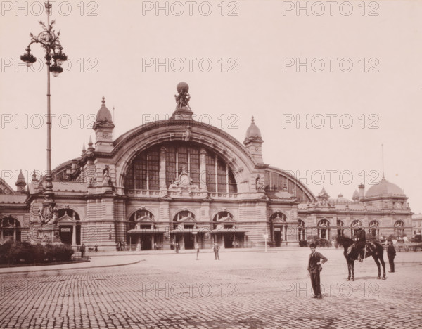 Equestrian statue on a pedestal of Emperor Wilhelm I, Frankfurt-am-Main, Germany, photograph, Germany. Sculptor was Clemens Buscher (1855–1916), who created the statue in 1896. It was destroyed and melted down during WWII., caption: 'Station Frankfurt', c. 1875 - c. 1900, photographer, paper, pasteboard, collotype, photogravure on photographic paper on cardboard, height 274 mm, width 202 mm x width 490 mm, height 330 mm, A large stone building with an ornate arched central facade and tall arched windows topped by sculpted figures and decorative carvings spans the scene, flanked by domed towers and smaller ornamental turrets; a tall ornate streetlamp rises to the left foreground, and a cobbled plaza stretches across the front. Several people stand near the building's entrances and in the plaza, and a man in a suit stands nearer to the camera with a horse and a handler slightly behind him to the right. The sky is empty above the building, and architectural details such as columns, statue...