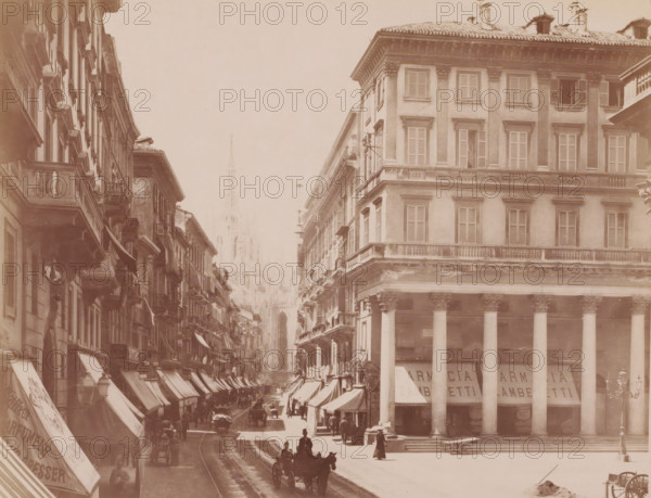Corso Vittorio Emanuele, Milan, Italy, photograph, View of the street Corso Vittorio Emanuele with shop fronts, horse and carriages and pedestrians, Italy., caption: '6830a MILANO. Corso Vittorio Emanuele.', caption: '(EDIZIONI BROGI)', photographer: Edizioni Brogi, Florence, publisher: Giacomo Brogi, c. 1860 - c. 1890, Tuscany, pasteboard, paper, albumen print, height 197 mm x width 249 mm, height 492 mm x width 330 mm, Lombardy, A sepia-toned street scene showing tall multi-story buildings lining a narrow avenue, with rows of shuttered windows and ornate balconies, awnings extending over the sidewalks, and a colonnaded building with large columns on the right; a distant Gothic-style cathedral spire is visible through a light haze at the end of the street, while horse-drawn carriages and pedestrians occupy the roadway and sidewalks, tram rails running down the center, and street lamps and signage projecting from building facades.