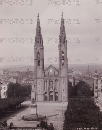 Katholische Hauptkirche', Wiesbaden, Germany, photograph, Frontal view of the church and the empty square in front of it from a high point of view., inscription: '127. Kat. Hauptkirche.', inscription: 'Mondel & Jacob Inh. G. Jacob, Hof-Phot. Wiesbaden 1896.', photographer: Mondel & Jacob, photographer: Mondel, photographer: Julius Jacob, 1896, Mondel & Jacob, Mondel, Julius Jacob, photographer, Hessen, pasteboard, paper, gelatin silver print, developing gelatin silver print, height 282 mm, width 225 mm x width 330 mm, height 508 mm, A large stone church façade with two tall ornate spires dominates the center of the image, each spire featuring intricate Gothic tracery and pointed finials, and the central section between them showing a triangular gable with a circular clock and a cross at its peak; below the gable are tall arched doorways and detailed sculptural relief above the main entrance. In front of the church is an open square with a tall obelisk monument on a stepped base enclose...
