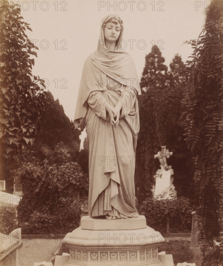 Furnaces of the Crematorium, Cimitero Monumentale, Milan, Italy, photograph, Italy., caption: '(Edizioni Brogi)', caption: '960. MILANO - Cimitero Monumentale - Forni del Crematoio', photographer: Edizioni Brogi, Florence, publisher: Giacomo Brogi, c. 1860 - c. 1890, Tuscany, pasteboard, paper, albumen print, height 250 mm, width 203 mm x width 510 mm, height 330 mm, Lombardy, A pale stone statue of a robed female figure stands on an ornate pedestal, the figure wearing a draped hood and long flowing garments gathered at the waist with hands gently clasped at waist level and toes visible at the hem, flanked by tall leafy plants and dark conical trees in the background with a partially visible decorative cross-shaped monument among the foliage.