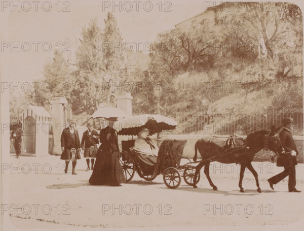Queen Victoria in her carriage in a park, France ?, photograph, with staff and two guards., signature: '... COUTON, NICE', caption, photographer: Claudius Couton, Nice, c. 1890 - 1901-01-22, Provence-Alpes-Côte d'Azur, paper, pasteboard, daylight gelatin silver print, daylight gelatin silver print on photo paper on cardboard, height 175 mm x width 229 mm, height 303 mm x width 388 mm, A sepia-toned street scene showing a horse-drawn carriage with a scalloped canopy holding a seated woman wrapped in a shawl, a standing woman beside the carriage holding an umbrella, two men walking nearby in formal attire with long coats and hats, a man leading the horse at the front of the carriage, a gated fence and lamp post behind them, a small guardhouse or sentry box to the left, and a hillside covered with trees and vegetation in the background.