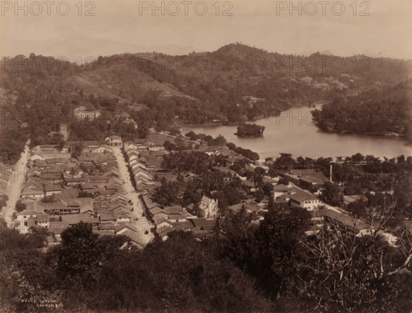 View of Kandy, Sri Lanka, photograph, View of the city of Kandy in the district of the same name, Sri Lanka., inscription: 'Kandy, Ceylon., Scowen & Co.', Ceylon', photographer: Charles T. Scowen & Co., Kandy, c. 1870 - c. 1890, Charles T. Scowen & Co., photographer, South Asia, pasteboard, paper, albumen print, height 213 mm, width 281 mm x width 407 mm, height 323 mm, A sepia aerial view showing a town with rows of tiled roofs and narrow streets leading toward a waterfront, a river or lake curving through the middle ground with a small vegetated island near its center, tree-lined banks and scattered buildings along the shoreline, clusters of trees and palms interspersed among houses, a prominent church-like building with a pointed roof near the street, larger structures set back amid greenery, a wooded foreground with dense foliage, and rolling forested hills rising in the background under a hazy sky.
